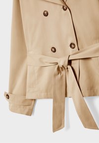 Béžový trenčkot s dvojitým zapínáním, velkými hnědými knoflíky a páskem v pase. Vyznačuje se hladkou texturou a jemným leskem.