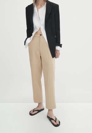 Kvinde iført beige højtaljede bukser, hvid knappet skjorte delvist ude af bukserne, sort dobbeltradet blazer og sorte flipflops.