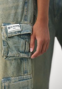 Lys blå denim overalls med en frontlomme, falmet tekstur, hvit etikett som sier "BDG JEANS," og hånden hvilende på kanten av lommen.