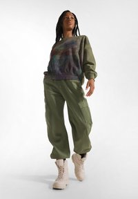 Olivegrüner übergroßer Pullover mit buntem abstraktem Print; kombiniert mit olivfarbenen Cargo-Hosen mit Seitentaschen; hellbeige Kampfstiefeln.