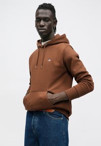 Barna kapucnis pulóver elülső zsebbel, húzózsinóros kapucnival és hímzett logóval. Kék farmernadrággal párosítva. Laza, kényelmes fazon.