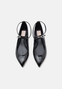 Chaussures plates noires en cuir verni à bout pointu avec brides à la cheville et boucles argentées, vues de dessus sur un fond blanc.