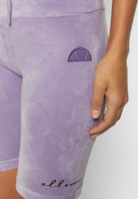 Gros plan sur une main posée sur un short en velours lavande avec taille à cordon de serrage et petit logo semi-circulaire violet au-dessus d'un texte noir brodé.