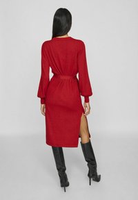 Rode, langmouwige gebreide jurk met een ceintuur op de taille en een zijnaad; gecombineerd met kniehoge zwarte laarzen; gladde textuur en relaxte silhouet.