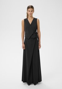 Vrouw in een mouwloos zwart wikkelvest met gesp en bijpassende lange wijde broek, staand tegen een effen witte achtergrond.