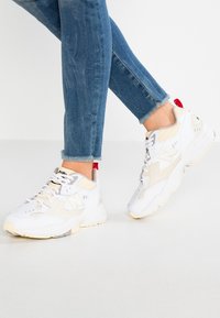 Baskets blanches et beiges avec un design texturé, dotées d'un laçage à l'avant, d'un accent rouge au talon et d'une semelle en caoutchouc épaisse.