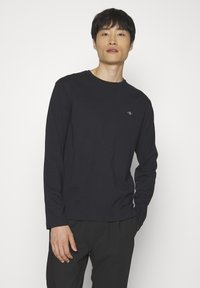 Camiseta negra de manga larga hecha de tela suave, con cuello redondo y un pequeño logotipo en el pecho. Diseño sencillo y ajustado.