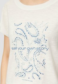 Weißes Baumwoll-T-Shirt mit kurzen Ärmeln, das blaue abstrakte Muster und den Text "Erzähle deine eigene Geschichte" in einer fetten Schriftart zeigt.