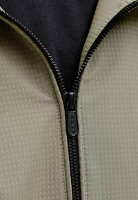 Detailný pohľad na čiastočne zapnutý čierny zips na textúrovanej svetlozelenej bunde na pozadí z čiernej látky.