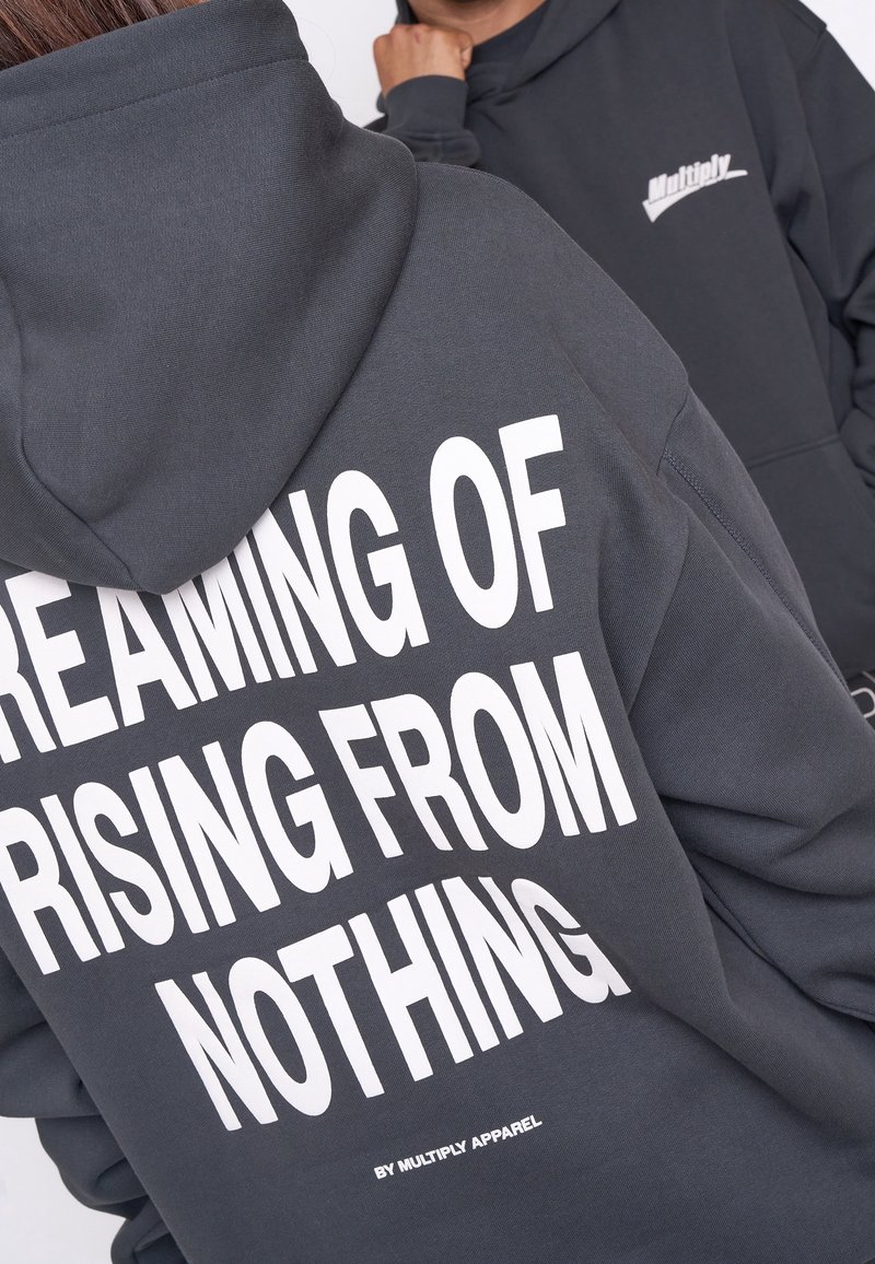 Sudadera gris de mezcla de algodón con un audaz texto blanco: "SOÑANDO CON ELEVARSE DESDE NADA" y "POR MULTIPLY APPAREL" en la parte inferior.