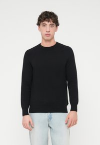 Μαύρο πλεκτό πουλόβερ με μακριά μανίκια, ριμπ λεπτομέρειες στο ντεκολτέ και τις μανσέτες, και άνετη εφαρμογή. Φορεμένο με ανοιχτό μπλε τζιν.