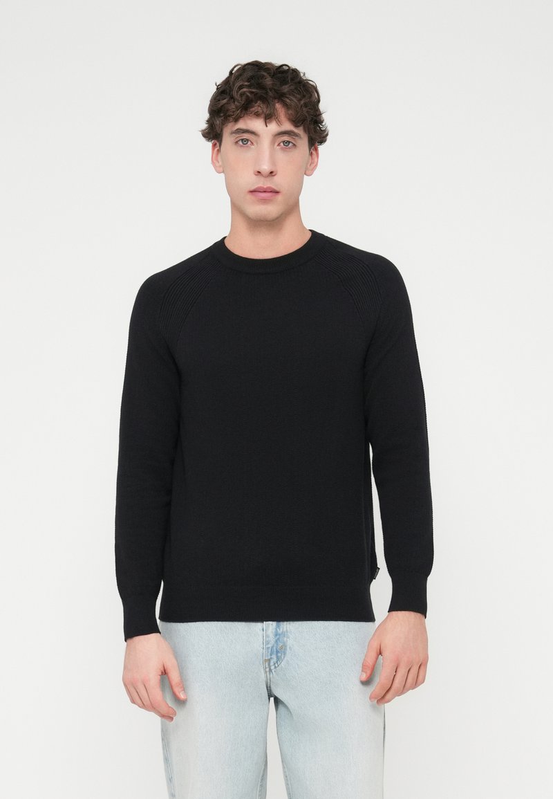 Μαύρο πλεκτό πουλόβερ με μακριά μανίκια, ριμπ λεπτομέρειες στο ντεκολτέ και τις μανσέτες, και άνετη εφαρμογή. Φορεμένο με ανοιχτό μπλε τζιν.