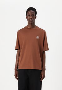 Καφέ βαμβακερό μπλουζάκι με άνετη εφαρμογή, κοντά μανίκια και στρογγυλή λαιμόκοψη. Διαθέτει ένα μικρό ε�κτυπωμένο γραφικό στην αριστερή πλευρά του στήθους.