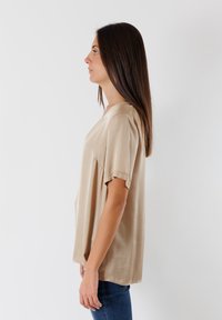 Top beige a maniche corte con vestibilità ampia, scollatura a girocollo e un leggero luccichio. Il tessuto appare liscio con una morbida caduta ai lati.