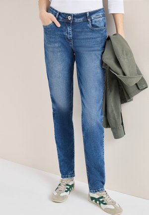Personne portant un jean slim bleu et des baskets blanches, tenant une veste verte pliée dans une main, debout contre un mur beige.