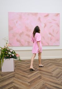Femme en robe rose marchant vers une grande peinture abstraite rose dans une galerie avec un sol en parquet à chevrons en bois et une composition florale à proximité.
