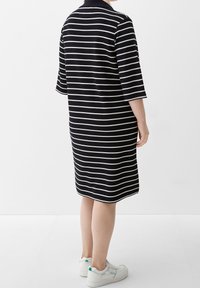 Persona in piedi, che indossa un vestito a righe orizzontali bianche e nere, lungo fino al ginocchio, con maniche a tre quarti e sneakers bianche.