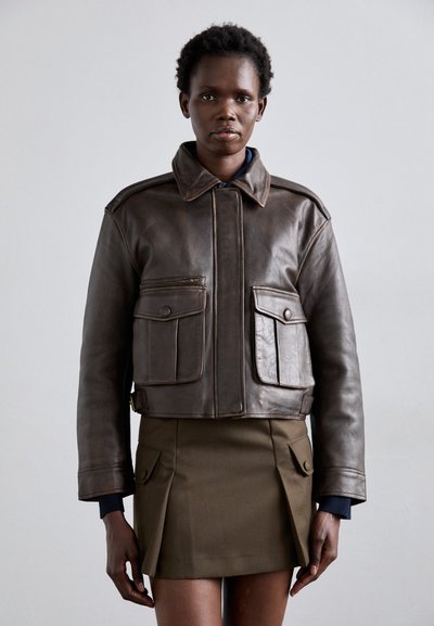 Giacca cropped in pelle marrone con colletto, due tasche frontali e cerniera laterale, abbinata a una gonna mini plissettata color khaki.