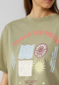 Olijf groene katoenen t-shirt met kleurrijke grafische prints, waaronder golven, een zon en tekst. Loose fit met een ronde halslijn en korte mouwen.