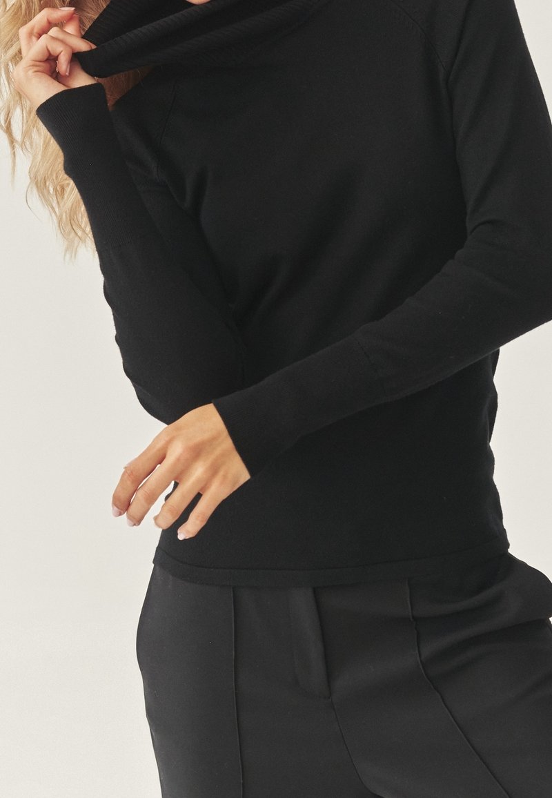 Femme ajustant le col d'un pull noir à col roulé à manches longues, portant un pantalon noir sur un fond clair uni.