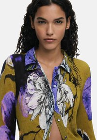 Camicia a bottoni a coste con un vivace motivo floreale in viola, nero e giallo. Il tessuto presenta un aspetto testurizzato con un dettaglio del colletto.