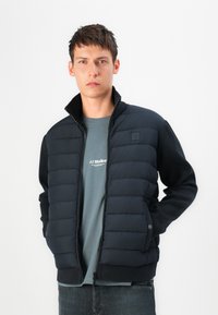 Black puffer jacket with a high collar, quilted front panels, and knit sleeves. Worn over a gray T-shirt with a small logo.
