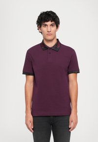 Jovem com cabelo encaracolado escuro, vestido com uma camisola polo ameixa com gola e punhos em preto e vermelho, de pé contra um fundo branco simples.