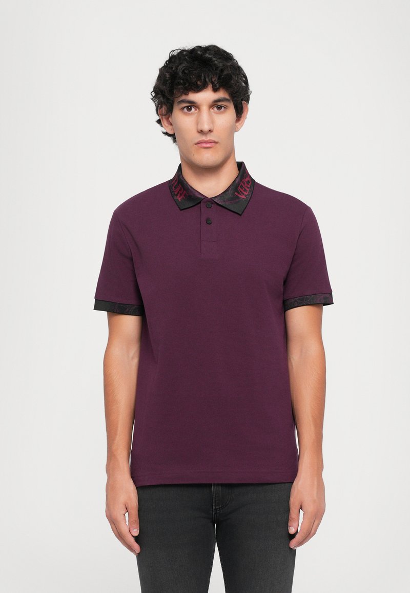 Jovem com cabelo encaracolado escuro, vestido com uma camisola polo ameixa com gola e punhos em preto e vermelho, de pé contra um fundo branco simples.