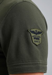 Camisa de manga corta verde oliva con un tejido texturizado, cuello redondeado y un parche bordado de estilo militar en la manga.