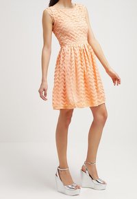 Robe de couleur pêche avec un motif en zigzag, sans manches, taille froncée et jupe évasée. Portée avec des sandales compensées argentées.