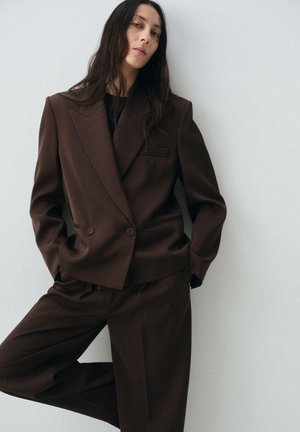 Costume marron sur mesure avec veste croisée et pantalon à jambes larges. Tissu lisse, revers nets et détail de poche poitrine unique.