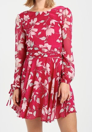 Femme portant une robe rouge à fleurs à manches longues, taille cintrée, jupe fluide et poignets noués, debout devant un fond uni.