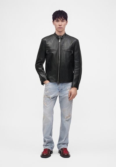 Homme portant une veste en cuir noire, un jean bleu clair déchiré et des bottes foncées avec des lacets rouges, debout devant un fond blanc uni.
