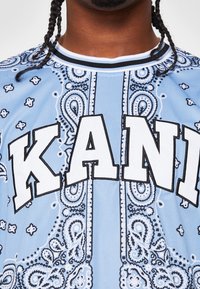 Světle modrá košile s krátkým rukávem, která má bílý vzor paisley a výrazný text "KANI" v černě obrysem vyvedených písmenech na přední straně.