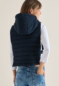 Marineblau gepolsterte Weste mit Kapuze, ausgestattet mit horizontaler Steppung und einem Frontreißverschluss. Getragen über einem weißen Langarmshirt und Jeans.