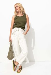 Haut sans manches olive, pantalon blanc taille haute avec liens à l'avant, sac à bandoulière gris et tongs noires avec accents verts.