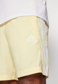 Ανοιχτό κίτρινες σορτς φτιαγμένες από μαλακό ύφασμα με άνετη εφαρμογή. Διαθέτουν λευκή τριπλή ρίγα και λευκό λογότυπο Adidas αριστερά.