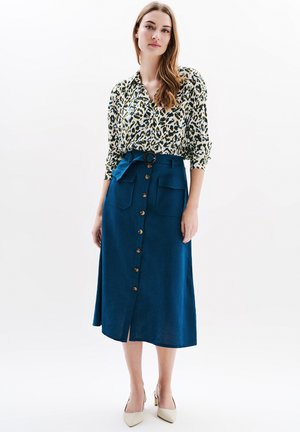 Chemise à manches longues à motif floral avec un design vert et blanc, associée à une jupe évasée bleue avec un bouton à l'avant et une taille à nouer.