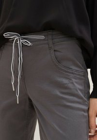 Pantaloni di cotone grigi con vita regolabile a cordino, caratterizzati da accenti bianchi e terminali argentati, insieme a tasche laterali e dettagli cuciti.