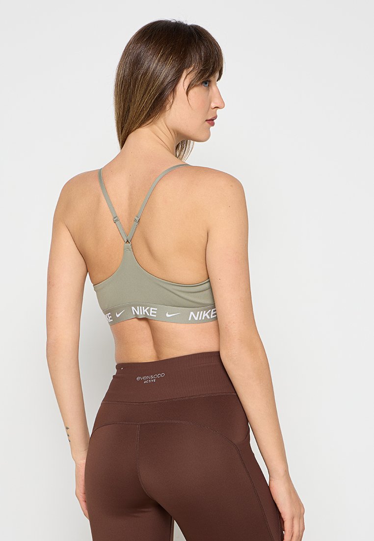Sujetador deportivo verde olivo con tirantes ajustables y una banda blanca con el logo de Nike. Combinado con leggins de talle alto color marrón oscuro. Textura suave.