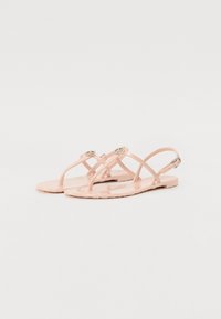 Sandales en jelly roses avec une semelle plate, dotées de brides croisées et d'accents métalliques ronds. Texture lisse avec une finition brillante.
