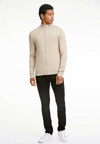 Beige zip-up tröja med hög krage, figurnära design, ribbade ärmslut, tillsammans med svarta slim-fit jeans och vita sneakers.