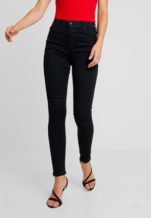 Femme portant un jean skinny noir, un top rouge sans manches et des sandales à talons noires à lanières, debout devant un fond gris.