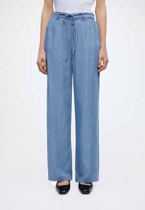 Άτομο που φοράει ανοιχτό μπλε παντελόνι με φαρδιά πόδια και κορδόνι, λευκό τοπ και μαύρα παπούτσια με τετράγωνη μύτη, στεκόμενο μπροστά σε απλό φόντο.