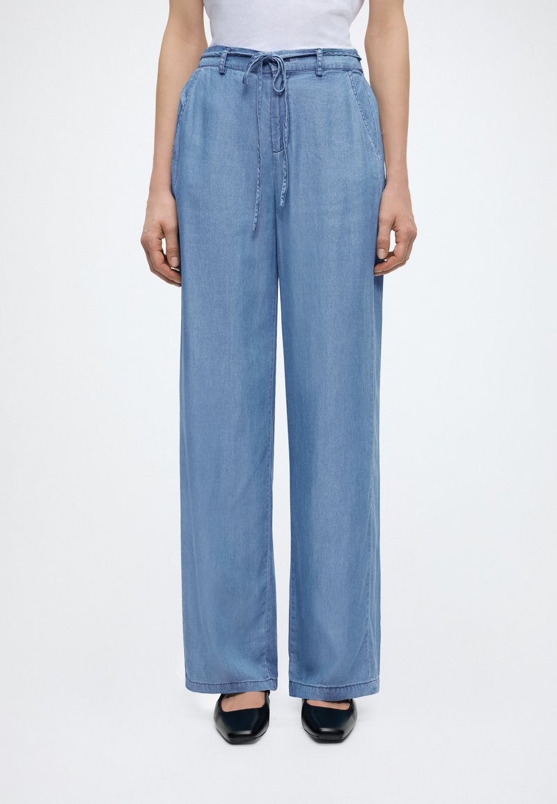 Άτομο που φοράει ανοιχτό μπλε παντελόνι με φαρδιά πόδια και κορδόνι, λευκό τοπ και μαύρα παπούτσια με τετράγωνη μύτη, στεκόμενο μπροστά σε απλό φόντο.