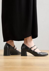 Scarpe con tacco in pelle nera con design a doppia cinghia e fibbie circolari. Tacco quadrato e superficie liscia. Posizionate accanto a una gonna nera.