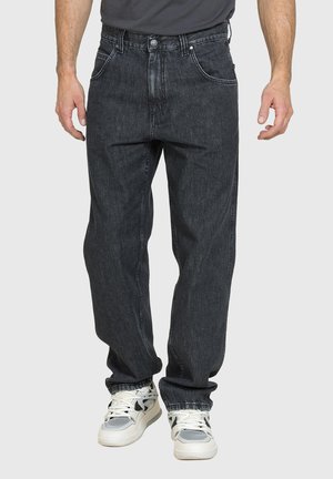 Uomo che indossa jeans larghi grigio scuro e sneakers bianche in piedi davanti a uno sfondo chiaro.