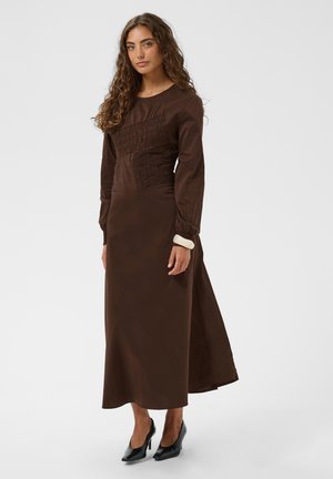 Femme aux longs cheveux bouclés portant une longue robe marron avec des panneaux texturés et des talons hauts noirs sur un fond gris clair.