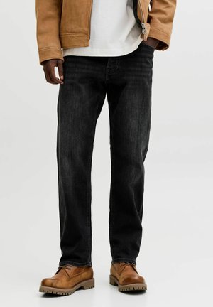 Mann trägt schwarze Jeans, braune Lederstiefel, weißes Hemd und braune Jacke, eine Hand in der Tasche, vor einfachem Hintergrund.