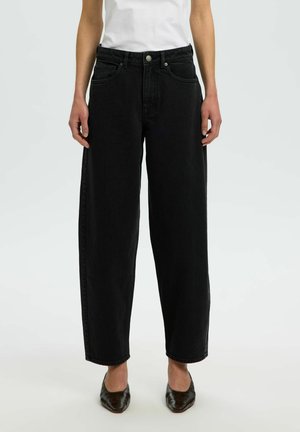 Personne portant un jean noir taille haute à jambes larges, associé à des chaussures noires à bout pointu et un haut blanc rentré.